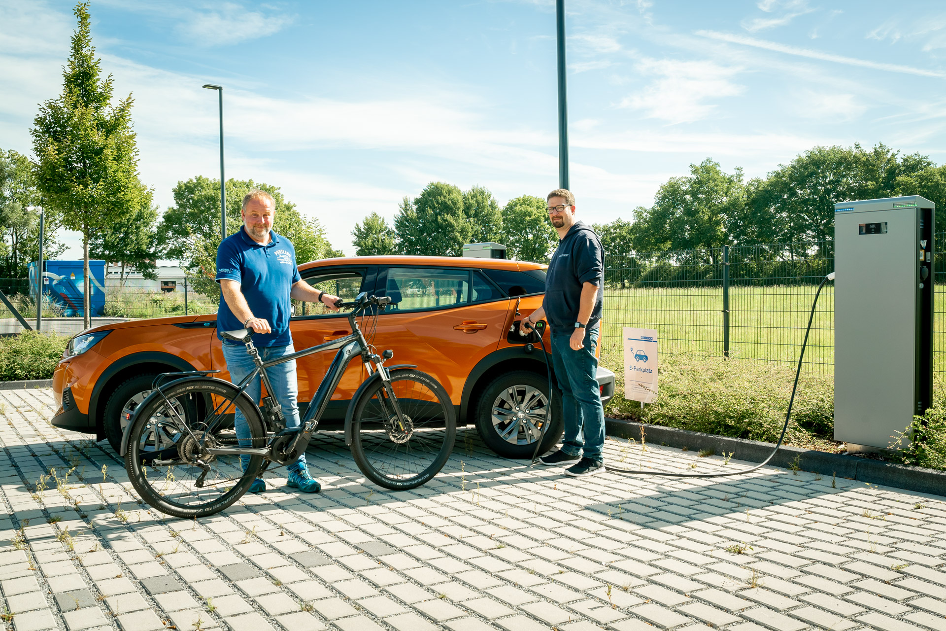 e-mobilitaet-fahrrad-leasing-weicon Zwei Männer stehen vor einem E-Auto und laden es auf, der linke hält ein Fahrrad in der Hand